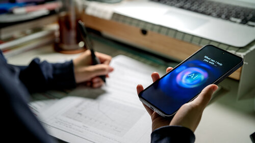 Ein Student kommuniziert über sein Smartphone mit einem KI-Chatbot, während er an einem Schreibtisch mit Laptop, Notizen und Schreibutensilien lernt.